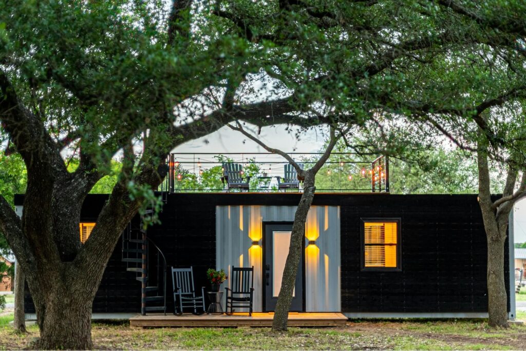 A single-story container home with black exterior surrounded by trees