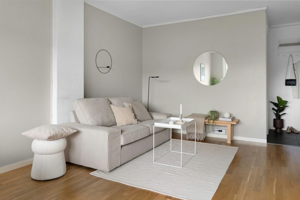 Minimalist living room with light beige tones. Features a sofa, a round wall mirror, a floor lamp, and a small table on a rug. Cozy and serene ambience.